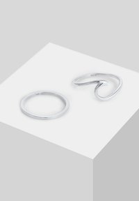Două inele argintii expuse pe o suprafață albă: un band subțire circular și un design fluid, în buclă, cu un finisaj lustruit.