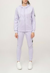 Sweat à capuche lavande claire avec fermeture éclair devant et deux poches, associé à un jogging ajusté assorti avec une poche latérale et des baskets blanches.