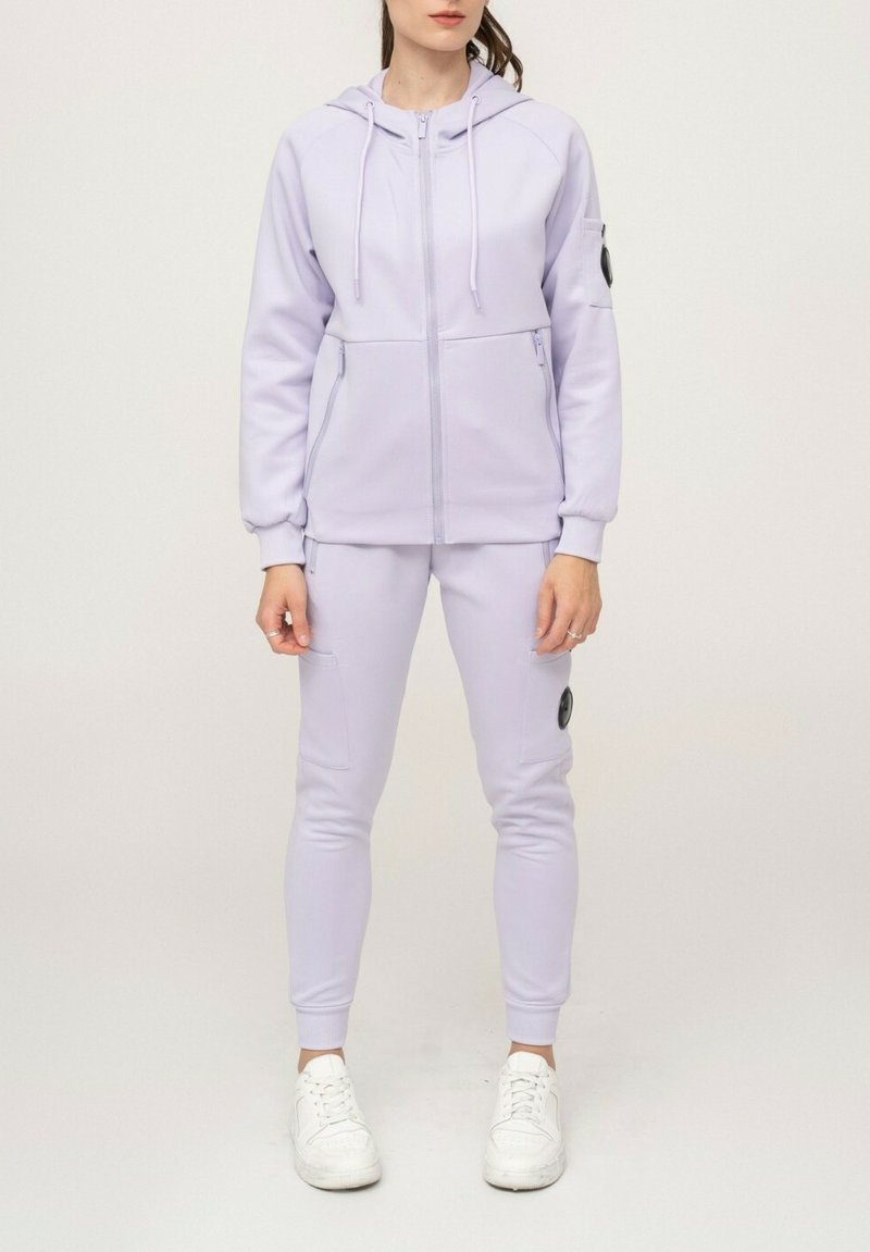 Sweat à capuche lavande claire avec fermeture éclair devant et deux poches, associé à un jogging ajusté assorti avec une poche latérale et des baskets blanches.