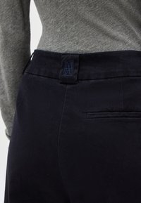 Κοντινό πλάνο ενός ατόμου που φοράει σκούρο μπλε παντελόνι και γκρι πλεκτό τοπ, εμφανίζοντας την πίσω ζώνη μέσης με ραμμένο λογότυπο.