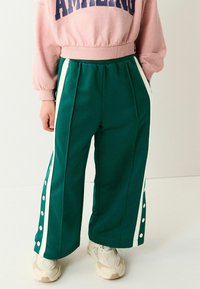 Pantalons de sport verts avec des rayures blanches sur les côtés et des boutons-pression ; tissu doux et texturé ; coupe ample ; associés à des baskets beiges.