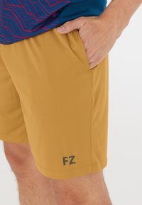 Ljust bruna idrottsshorts gjorda av mjukt tyg, med sidofickor och en diskret "FZ"-logotyp i mörkgrått nära nederkanten.