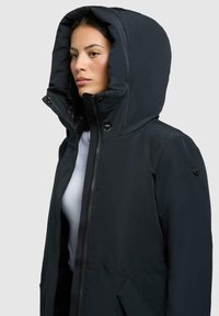 Schwarze Kapuzenjacke mit hohem Kragen, ausgestattet mit einem Frontreißverschluss, Seitentaschen und einem strukturierten Stoff. Die Kapuze ist groß und plüschig.