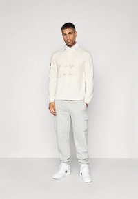 Mand står mod en ensfarvet baggrund iført en cremet langærmet skjorte, lysegrå cargo-sweatpants og hvide høj-top sneakers.