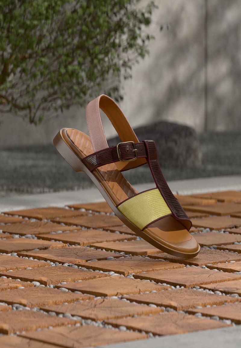 Καφέ και κίτρινο επίπεδο σανδάλι με λουράκι σε σχήμα Τ και αγκράφα στον αστράγαλο, τοποθετημένο πάνω σε υφή ξύλινης πλακίδας σε εξωτερικό χώρο με θολό φόντο από πράσινα φυτά.