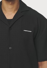Czarna koszula z krótkim rękawem o prążkowanej fakturze, z kołnierzem z wycięciem i zapięciem na guziki z przodu, z logo "PEGADOR" na lewej piersi.