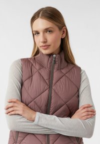 Steppweste in Mauve mit hohem Rippenkragen, zentrale Reißverschluss und einer leichten, glänzenden Textur. Darunter ein langärmliges graues Hemd.