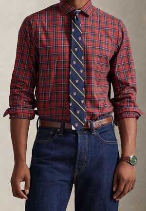 Mężczyzna w podwiniętej czerwonej koszuli w kratę, granatowym krawacie w żółte paski z herbem, ciemnoniebieskich dżinsach, brązowym skórzanym pasku i zegarku.