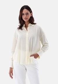 Camicia gialla chiaro con pois testurizzati, maniche lunghe e vestibilità rilassata. Abbinata a pantaloni bianchi con orlo leggero.
