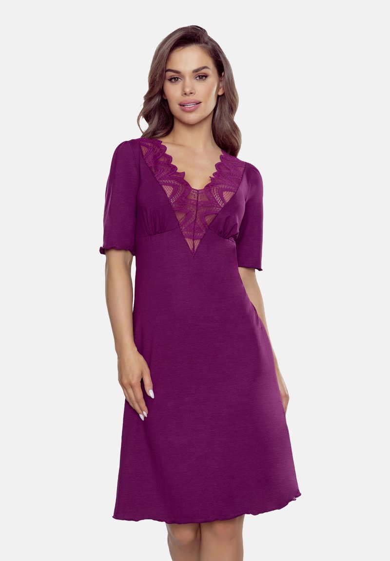 Modelka ubrana w purpurową sukienkę z koronkowym wykończeniem, z krótkimi rękawami i dekoltem w kształcie litery V, stojąca na jednolitym tle.