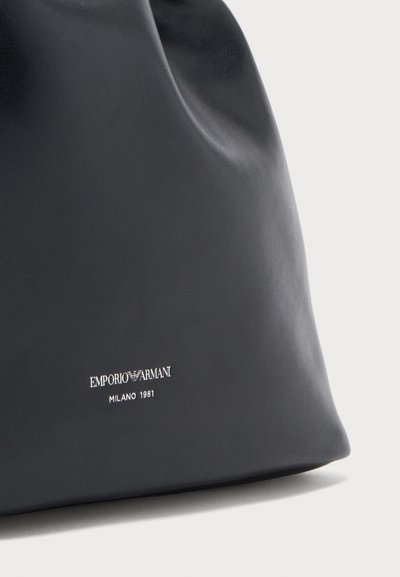 Μαύρη δερμάτινη γυναικεία τσάντα Emporio Armani με ασημένιο λογότυπο και το κείμενο "Milano 1981" σε λεία επιφάνεια, μπροστά σε απλό φόντο.