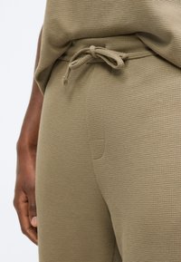 Olívazöld, texturált pamutnadrág húzózsinóros derékrésszel, kis zsebbel az oldalon, és közelkép a szövet mintájáról.