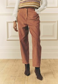 Pruunid veluurriidest püksid sirge lõikega, kõrge vöökohaga ja peene soonelise tekstuuriga, mida kannavad Burgundia värvi teravnokkade kõrged kontsad.