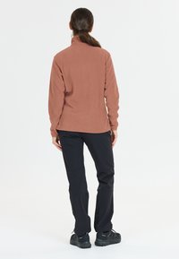 Kvinna med brun hästsvans som bär en brun fleecejacka, svarta byxor och svarta vandringsskor, stående med ryggen vänd mot en vit bakgrund.