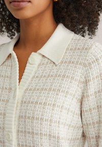 Ľahký krémový sveter na gombíky s textúrovaným vzorom, rebrovaným golierom a zapínaním na gombíky. Vyznačuje sa jemným striedavým vzorom.