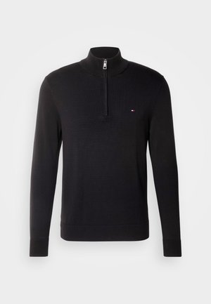 Juodas ilgas megotos megztinis su ketvirčio užtrauktuku ir mažas raudonas, baltas bei mėlynas logotipas kairėje krūtinės pusėje, pateiktas ant paprasto šviesiai pilko fono.