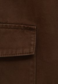 Gros plan sur un tissu marron avec des coutures détaillées le long du bord d'une poche à rabat rectangulaire.