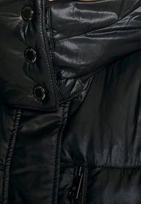 Svart vadderad jacka med en glansig finish, med stora svarta knappar, ficka med dragkedja och kviltad textur. Knappad krage synlig.