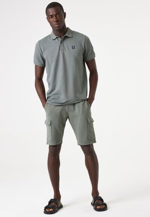 Polo verde con un motivo testurizzato, abbinato a pantaloni cargo coordinati con tasche laterali e sandali neri con design a fascia.