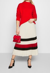Suéter de punto rojo, falda plisada crema con rayas negras y rojas, combinada con un pequeño bolso rojo y zapatos de tacón negros.