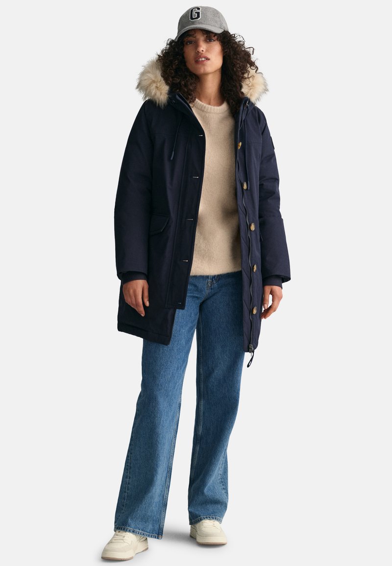 Marine Parka mit Kunstfellkapuze, ausgestattet mit Knöpfen und Fronttaschen. Kombiniert mit einem beigen Pullover und ausgestellten Jeans. Helle Sneakers.