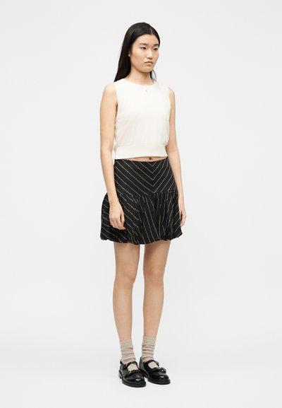 Γυναίκα που στέκεται φορώντας λευκό αμάνικο crop top, μαύρη φούστα με λευκές διαγώνιες ρίγες, μπεζ κάλτσες και μαύρα παπούτσια, μπροστά σε απλό φόντο.