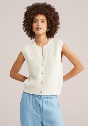 Ženska s kodrastimi lasmi, oblečena v belo pleteno telovnik brez rokavov z gumbi in svetlo modre hlače z vrvico, stoji z rokami na bokih.
