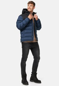 Mørkeblå og sort polstret jakke med hætte, der har lynlås lukning og ribbede manchetter. Båret over en beige bluse og parret med mørke jeans og støvler.