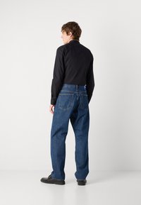 Sort langærmet skjorte, denimblå vidde jeans og sorte sko. Synlige syninger og lomme detaljer på jeansene.
