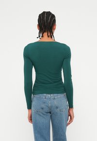 Langärmliges grünes Oberteil mit figurbetontem Design und geraffter Textur am Rücken, kombiniert mit hellblauen Jeans. Rückansicht dargestellt.