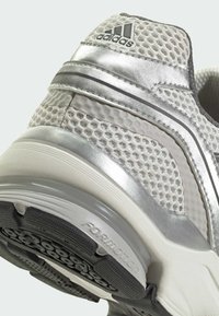 Srebrno-szary but sportowy z siateczkową cholewką, odblaskowymi akcentami i czarną gumową podeszwą z teksturowanym wzorem zapewniającym przyczepność. Obecne oznaczenia marki.