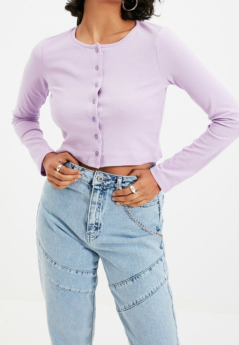 Lilla pitkähihainen nappipaita, jossa on pyöreä kaula-aukko ja teksturoitu, ribatoitu kangas. Yhdistetty vaaleansinisiin korkeavyötäröisiin farkkuihin, joissa on saumat.