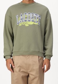 Oliwkowy sweatshirt z dużym logo "LACOSTE" w białym i żółtym kolorze, z ribbowanymi mankietami i dołem, wykonany z miękkiego materiału.