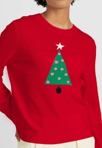 Roter gestrickter Pullover mit einem grünen Weihnachtsbaum, der pinke Punkte und einen weißen Stern an der Spitze trägt. Einfaches Design, leichtes Material.