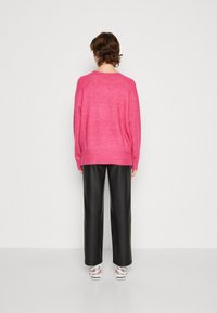 Osoba stojąca tyłem, ubrana w jasnoróżowy sweter, czarne szerokie spodnie oraz białe trampki, na jednolitym tle.