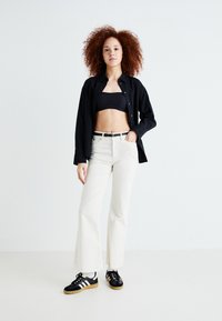 Utrekkende kremfargede bukser, svart cropped topp og en svart knappskjorte. Svart sneakers med hvite striper og gummisåle. Krøllete hår, nøytral bakgrunn.