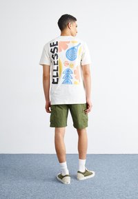 White t-shirt with colorful graphic design featuring a blue racquet, flowers, and abstract shapes, paired with green shorts and sneakers.