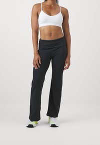 Biała sportowa biustonosz o cienkich ramiączkach, połączona z czarnymi spodniami dzwonowymi. Modelka nosi białe i zielone sneakersy, stojąc na jasnym tle.