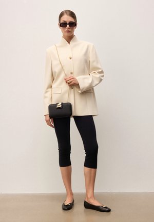 Femme portant un blazer crème oversize, des leggings noirs courts, des ballerines noires, des lunettes de soleil, et tenant un petit sac à main noir avec une bandoulière chaîne dorée.