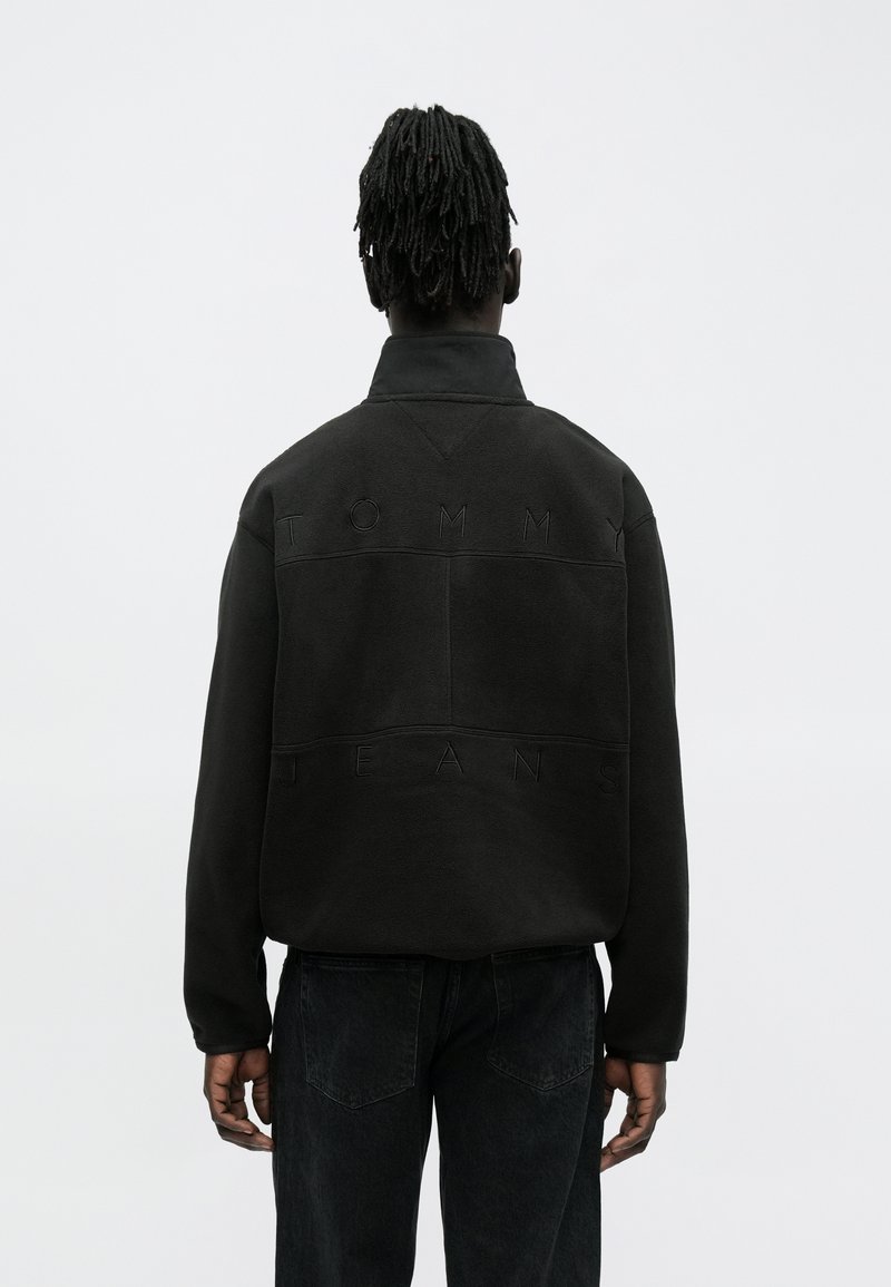 Μαύρο φλις πουλόβερ με ψηλό γιακά, με ανάγλυφη επιγραφή «TOMMY JEANS» στην πλάτη. Δομημένη, χαλαρή εφαρμογή με μακρυά μανίκια.