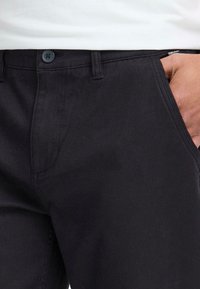 Close-up van zwarte broek met een knoop en zak, met een hand op de tailleband die de textuur en pasvorm benadrukt.