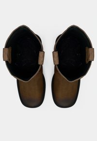 Bottes en cuir marron avec un bout rond et une semelle plate. L'intérieur est sombre, avec des accents en textile beige sur le haut.