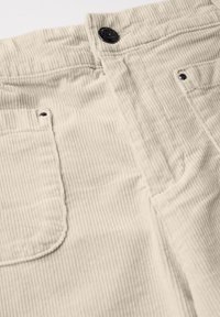 Beige corduroy broeken hebben verticale ribbels, een zwarte knoop in de taille en twee achterzakken met metalen accenten.