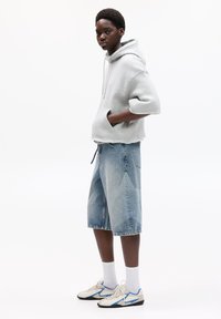 Haut cropped gris clair avec une poche avant. Short en denim bleu délavé avec un motif géométrique. Chaussettes blanches et baskets beige avec des accents bleus.