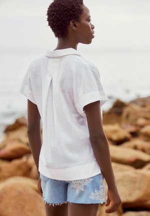 Chemise blanche à manches courtes en lin, dotée d'un pli au dos et d'une coupe décontractée, associée à un short en denim bleu clair avec des accents brodés.