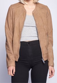 Braune durchbrochene Lederjacke mit Rundhalsausschnitt, Reißverschluss und schrägen Nähten. Getragen mit einem hellgrauen Oberteil und schwarzen Hosen.