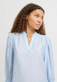 Blusa blu chiaro con una finitura testurizzata, dotata di scollatura a V e maniche a sbuffo, raccolte ai polsini. Strisce verticali sottili nel tessuto.