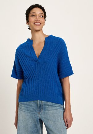 Woman smiling wearing a textured blue short-sleeve sweater with a split collar and light blue denim jeans against a plain background.