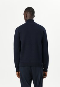 Ναυτικό πλεκτό πουλόβερ με ψηλό γιακά, ριμπ μανσέτες και στρίφωμα, και ομαλή υφή. Διαθέτει λεπτομέρειες στους ώμους για επιπλέον στυλ.