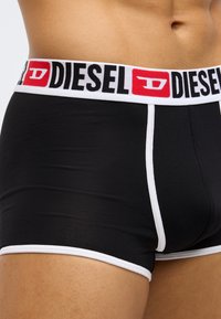Boxeri negri cu margine albă, cu o talie lată care afișează logo-ul „DIESEL” în roșu și negru. Textură de material neted.